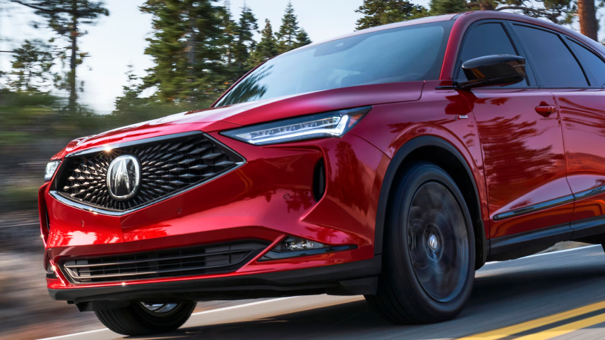 A medium shot of the front of an Acura driving on a road with trees in the background is shown. 
